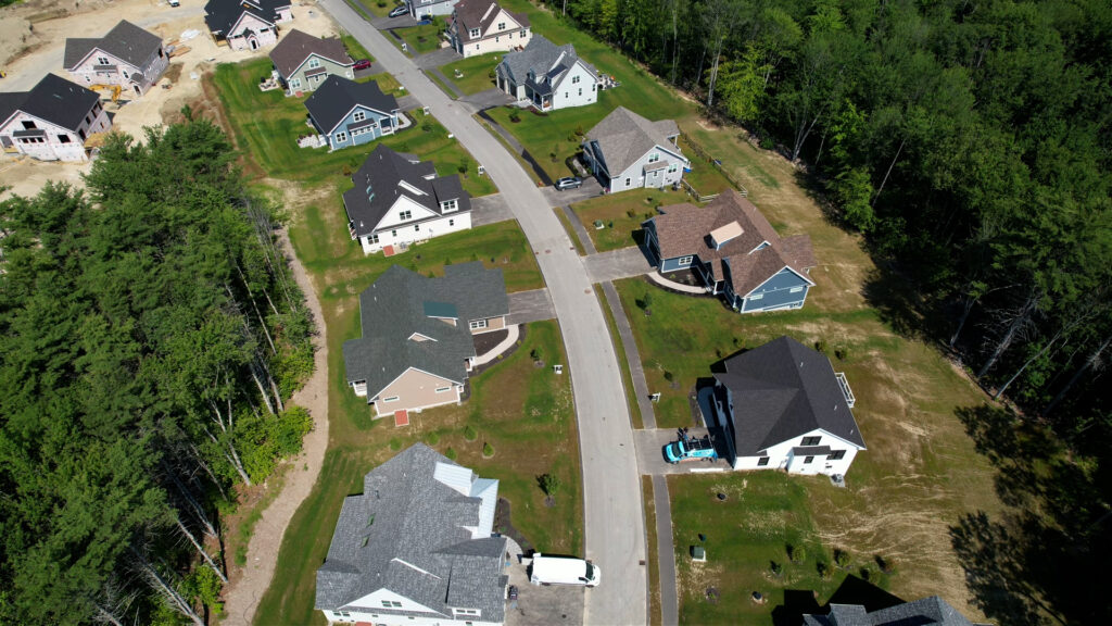 Aerial view of the Lady of the Lakes Estates neighborhood in Laconia, New Hampshire, with completed homes, new construction, landscaped yards, and surrounding woods.